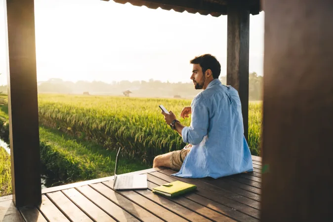 Seitenansicht eines konzentrierten jungen männlichen Freiberuflers im Hemd, der in der Nähe eines Laptops auf einem Holzpodest in einer malerischen Landschaft sitzt und bei Sonnenuntergang auf seinem Smartphone surft